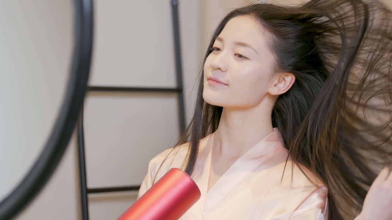 Una joven y hermosa mujer en albornoz sosteniendo y usando un secador de pelo eléctrico caliente en su largo cabello negro frente al espejo después de ducharse. Rutina matutina para cuidar la salud del cabello y empezar el día.