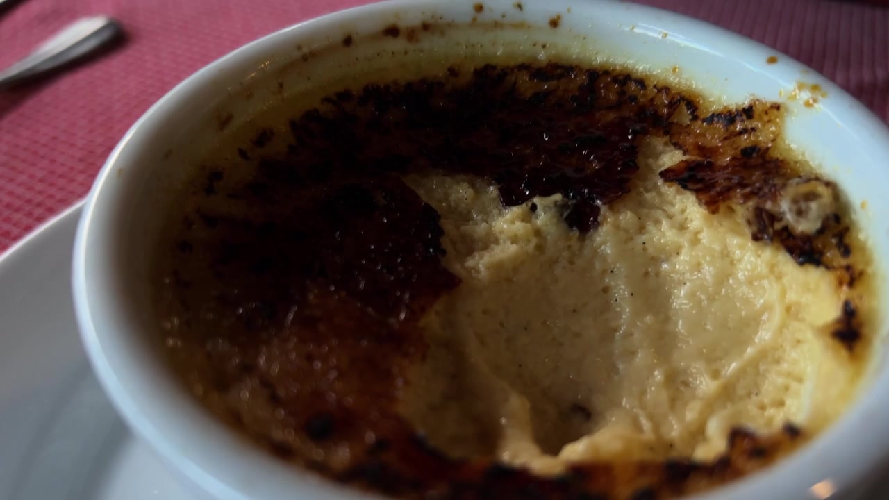 Vista cercana de una cuchara rompiendo la costra de azúcar caramelizada de un postre de crema catalana servido en un ramequín de cerámica en la mesa de un restaurante.