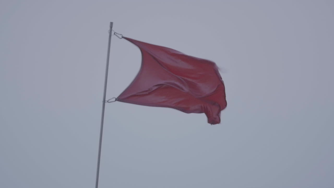 a Maroon Red flag waves in the grey skies. Blowing strongly in the wind, in slow motion.
