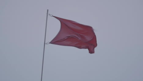 a maroon red flag waves in the grey skies. blowing strongly in the wind, in slow motion.