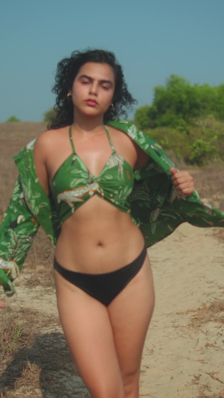 Donna sicura di sé che cammina su un sentiero sabbioso in bikini nero e top verde, giornata di sole.