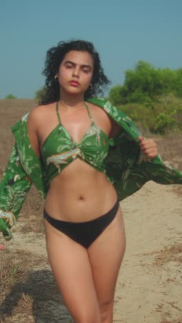 confident woman walking on sandy path in black bikini and green top, sunny day