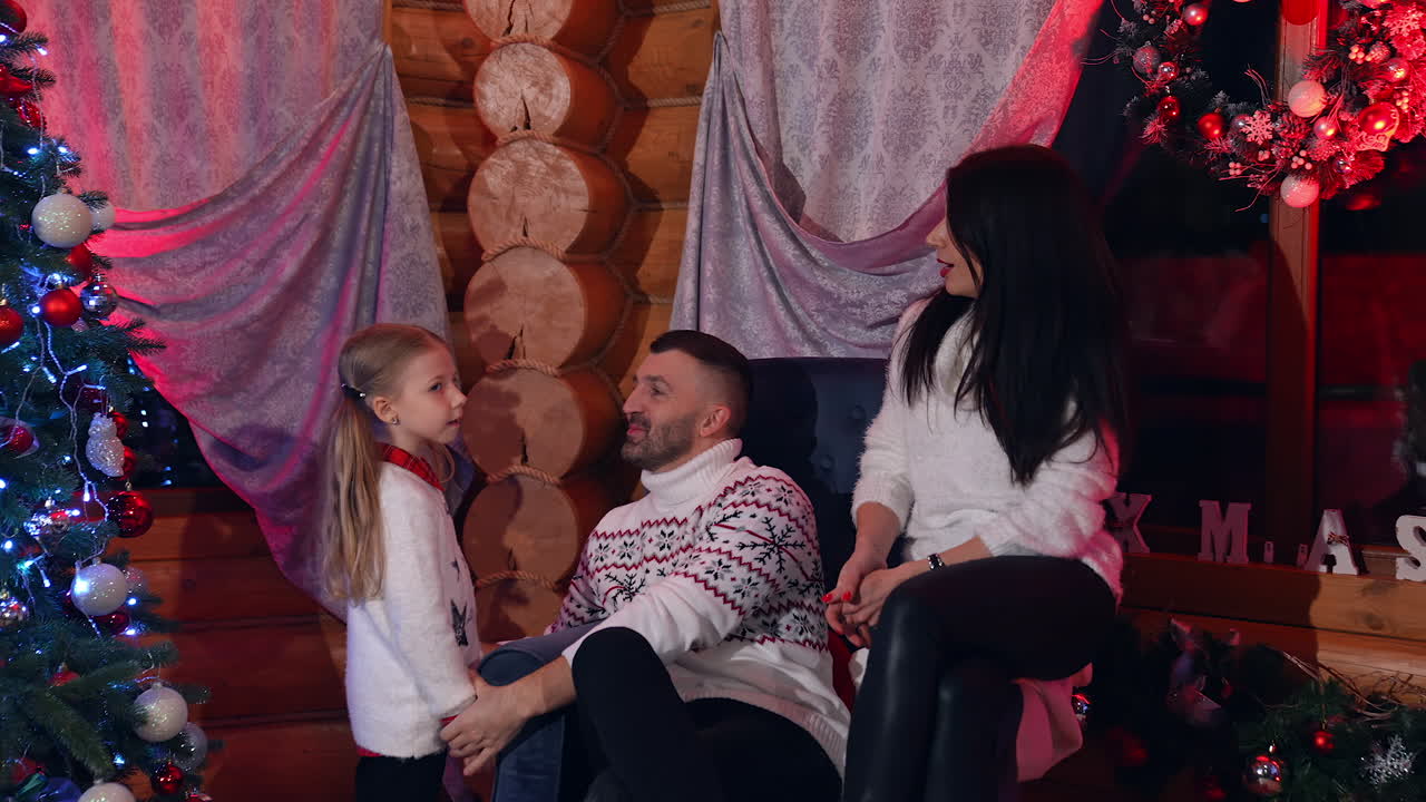 Happy parents sitting and chatting merrily in the comfortable sofa. Their little daughter comes to them and gives her drawing. Beautiful family near Christmas tree and decorative garlands.