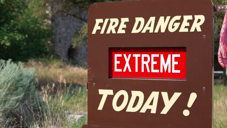 Insegna rossa e bianca con 'Pericolo estremo di incendio oggi' su un pannello metallico marrone, inquadratura ampia che si alza rapidamente per mostrare le montagne in un parco nazionale durante la crisi degli incendi boschivi negli Stati Uniti e nel mondo