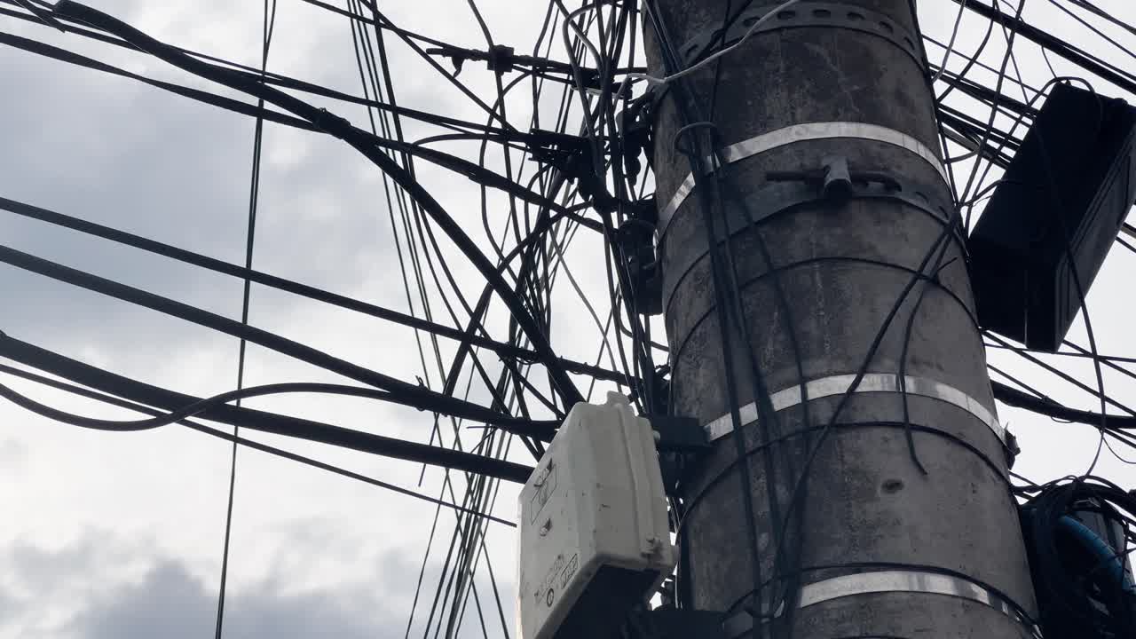 Pole in a poor neighborhood in Brazil, overloaded with tangled wires and apparent illegal electrical connections, commonly known as "gatos". The chaotic scene reflects urban infrastructure issues.