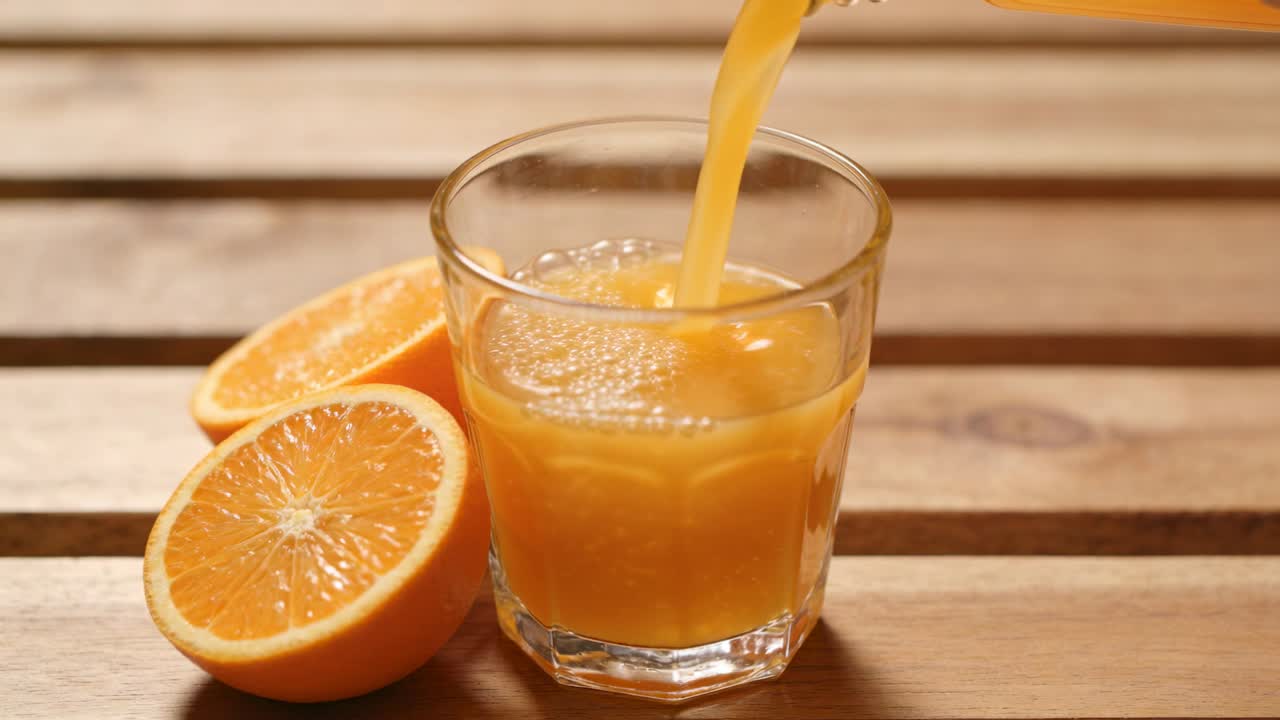 Kande vipper og hælder juice ned i klart glas på køkkenbord indtil næsten fuldt, med halverede appelsiner.