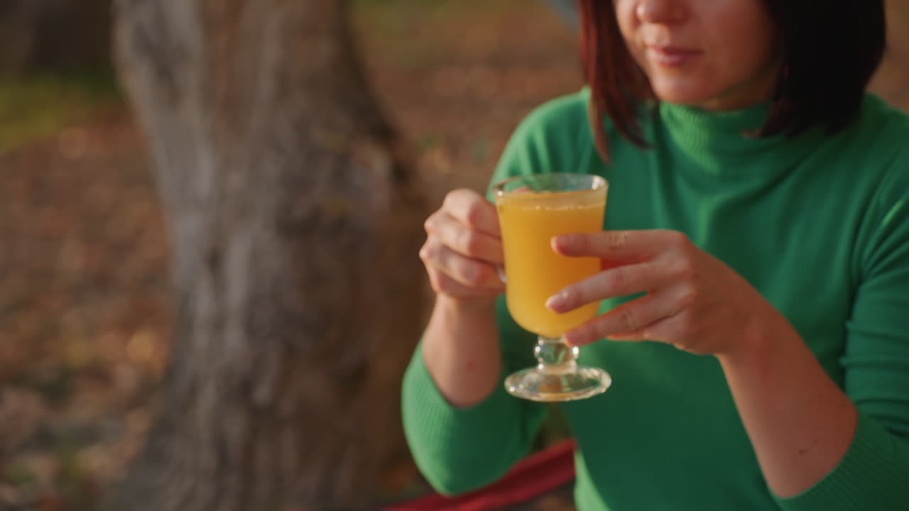 Campista mujer bebiendo zumo de naranja al aire libre, manos levantando el vaso del enfriador, sentada en una silla de camping roja junto a la hamaca, suéter verde y gafas de sol, desayuno tranquilo en un campamento de otoño y luz solar suave.