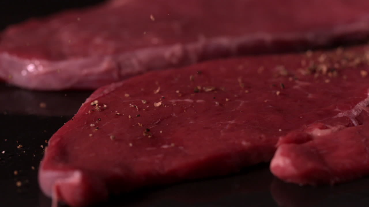 Beef steaks on black surface being seasoned