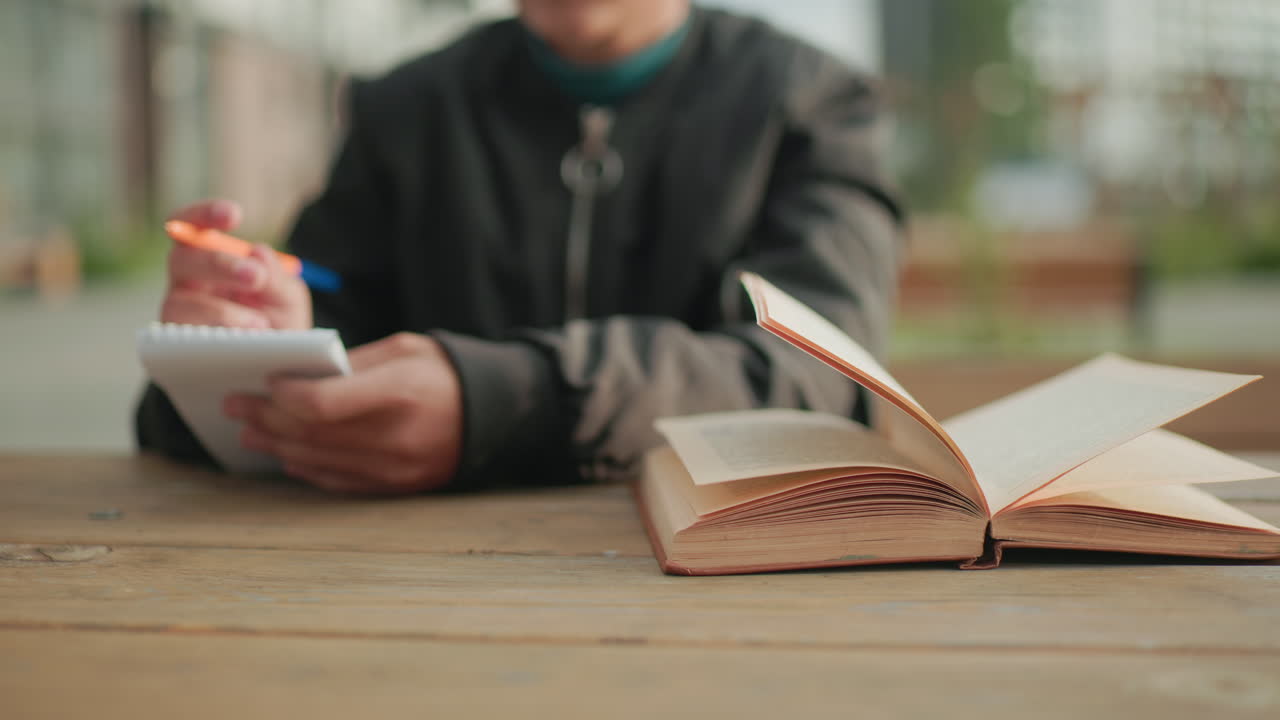 Close up of kid flipping to new page of open novel on wooden table outdoors with blurred bokeh background, pages lifting in breeze while holding notebook and pen