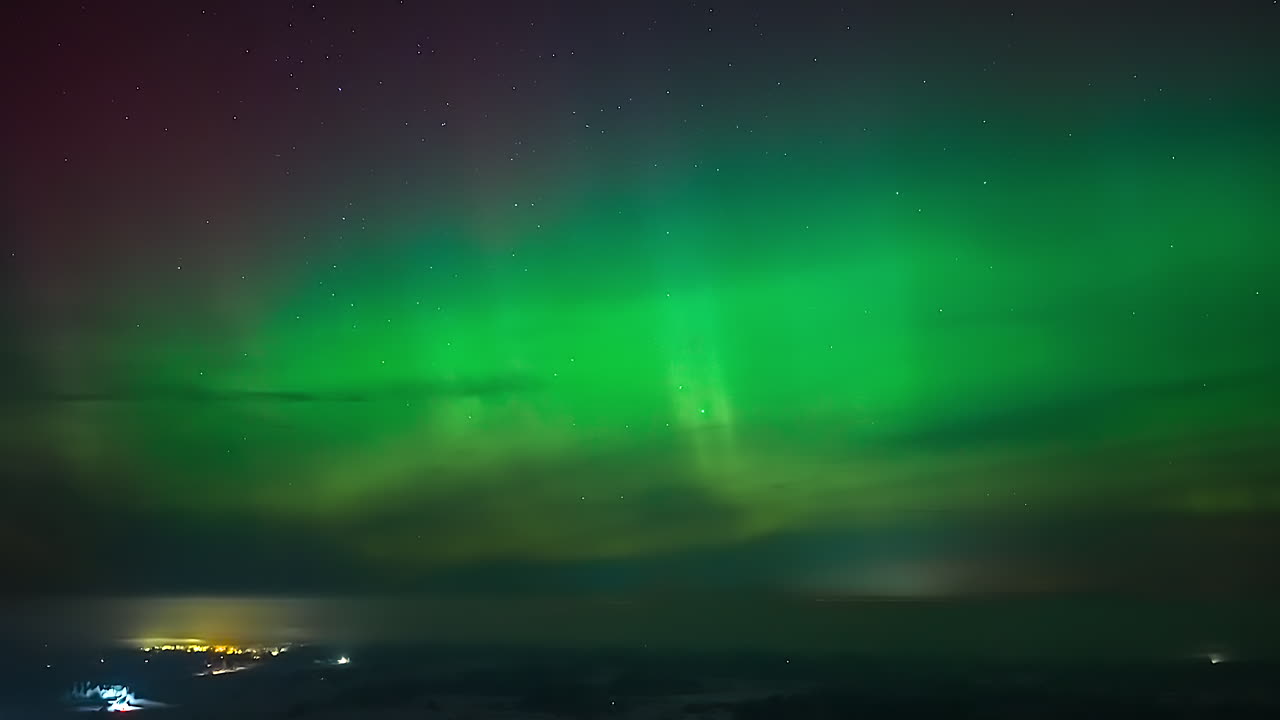 Timelapse dell'aurora boreale che risplende di verde con tonalità rosse sopra nuvole basse e un cielo notturno stellato, con un fioco bagliore della città vicino all'orizzonte.