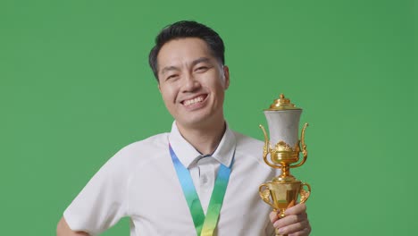 close up of asian man with a gold medal and trophy smiling to camera with one arm akimbo posing as the first winner on green screen background in the studio