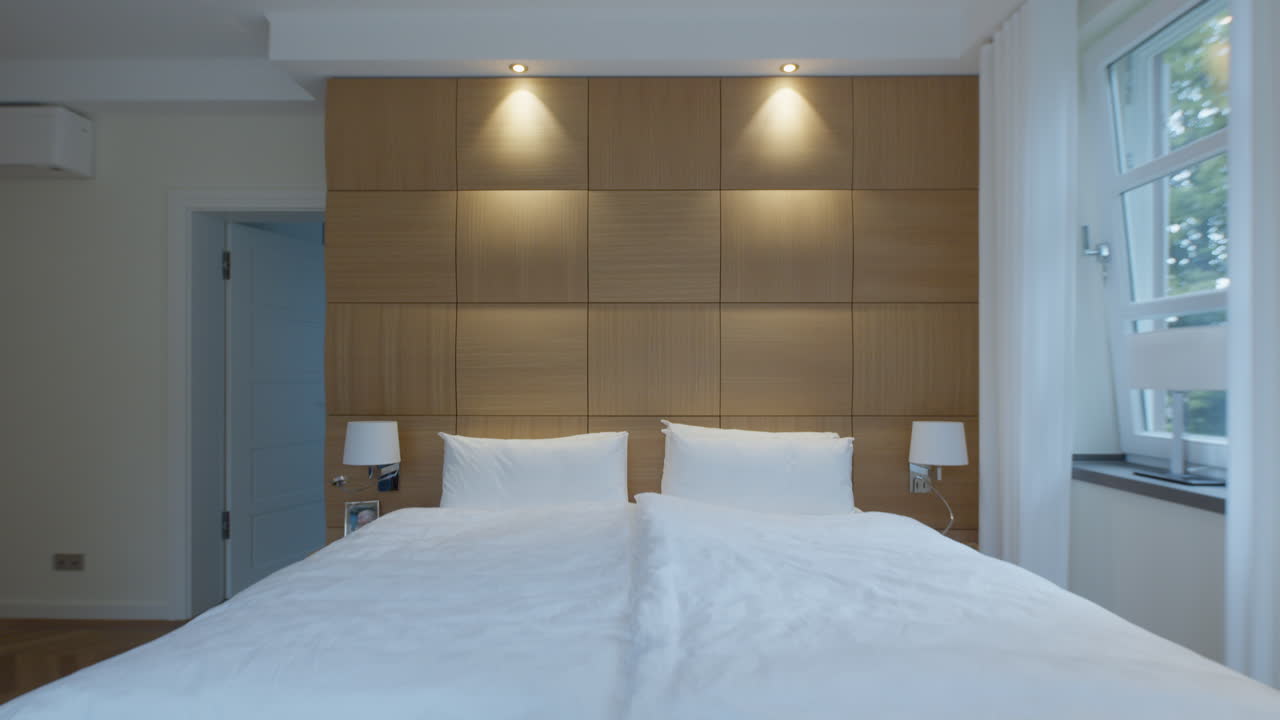High quality video of a cozy bedroom with soft wall lights, warm wood paneling and clean white bedding creating a calm and inviting atmosphere