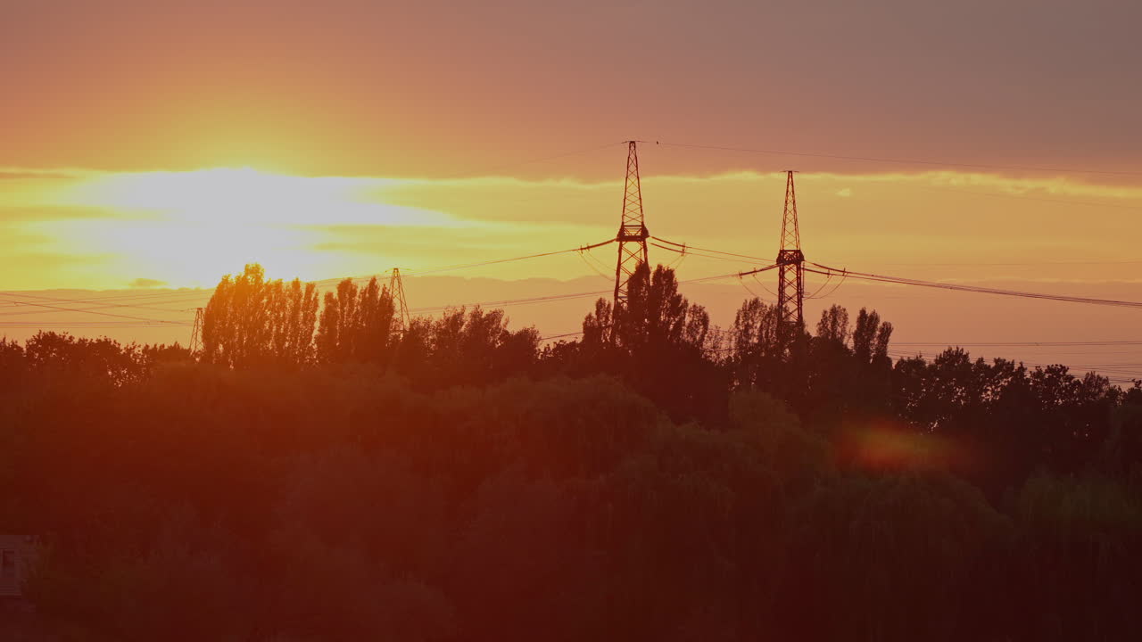 Tall electricity pylons at sunset. High voltage transmission towers and electric lines on the golden light. Panoramic view of sunset over the trees and electric towers in the evening.