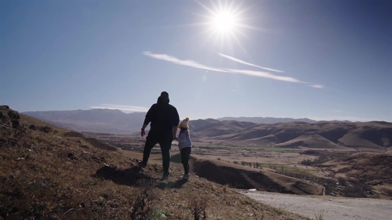 Um pai e uma filha caminham de mãos dadas por uma encosta ensolarada com vistas panorâmicas das montanhas em Tafí del Valle, Argentina, sob um céu claro de meio-dia.