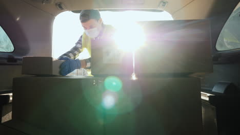 a young man in a medical mask loads boxes of medicines in the trunk of a car