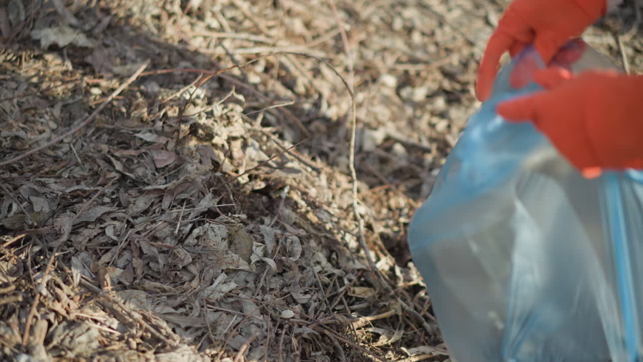 Persona che indossa guanti arancioni che mette rifiuti di plastica scartati in un sacco della spazzatura blu durante una pulizia all'aperto in un'area naturale secca, contribuendo alla protezione ambientale e alla riduzione dei rifiuti.