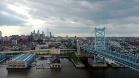 Luftdrohne,-Die-über-Den-Delaware-River-Fliegt,-Um-Die-Ben-Franklin-Bridge-Und-Die-Von-Sonne-Und-Wolken-Umgebene-Skyline-Von-Philadelphia-Zu-Zeigen