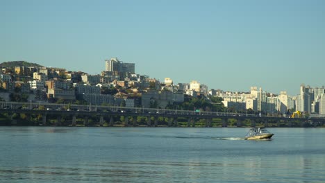 Small-boat-on-Hangang-or-Han-river-with-Yongsan,-Yongsan-gu-district-skyline-and-Gangbyeon-Expressway-on-background,-Seoul,-South-Korea