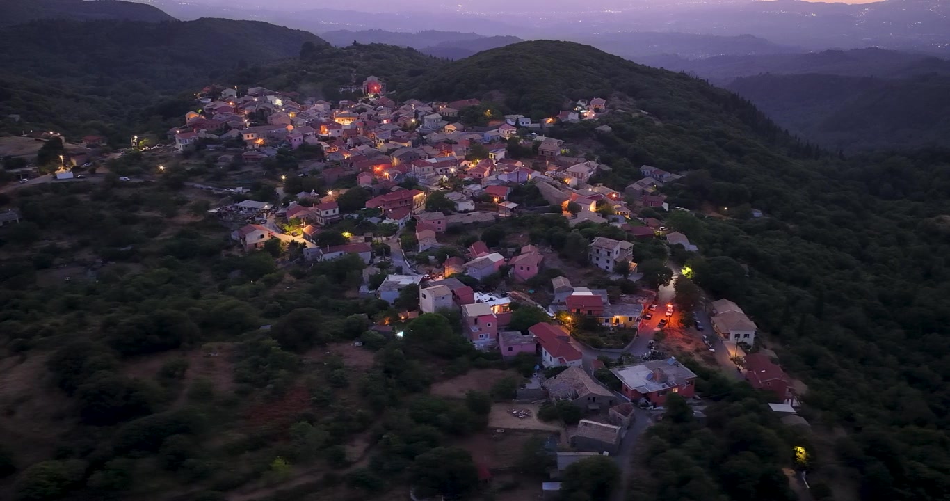 Ampia veduta aerea del tradizionale villaggio di Sokraki illuminato al crepuscolo a Corfù Nord, Grecia. Splendido paesaggio crepuscolare con case in pietra luminose circondate da lussureggianti colline montuose verdi.