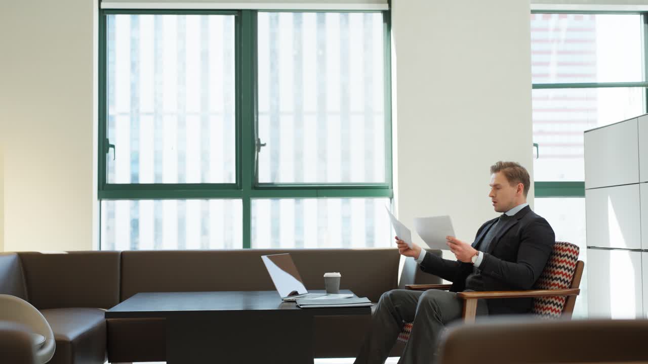 Angry stressed unsatisfied businessman in a suit throws documents papers in an office lounge with a laptop and coffee nearby - wide angle truck shot