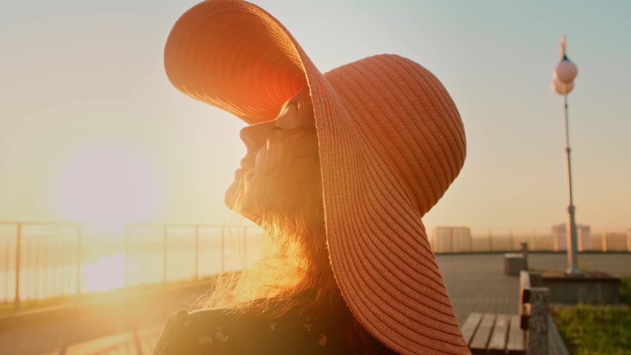 A Serene Sunset Scene with a Woman Wearing a Stylish Large Hat, Enjoying the Warm Glow of the Evening Light by the Waterfront