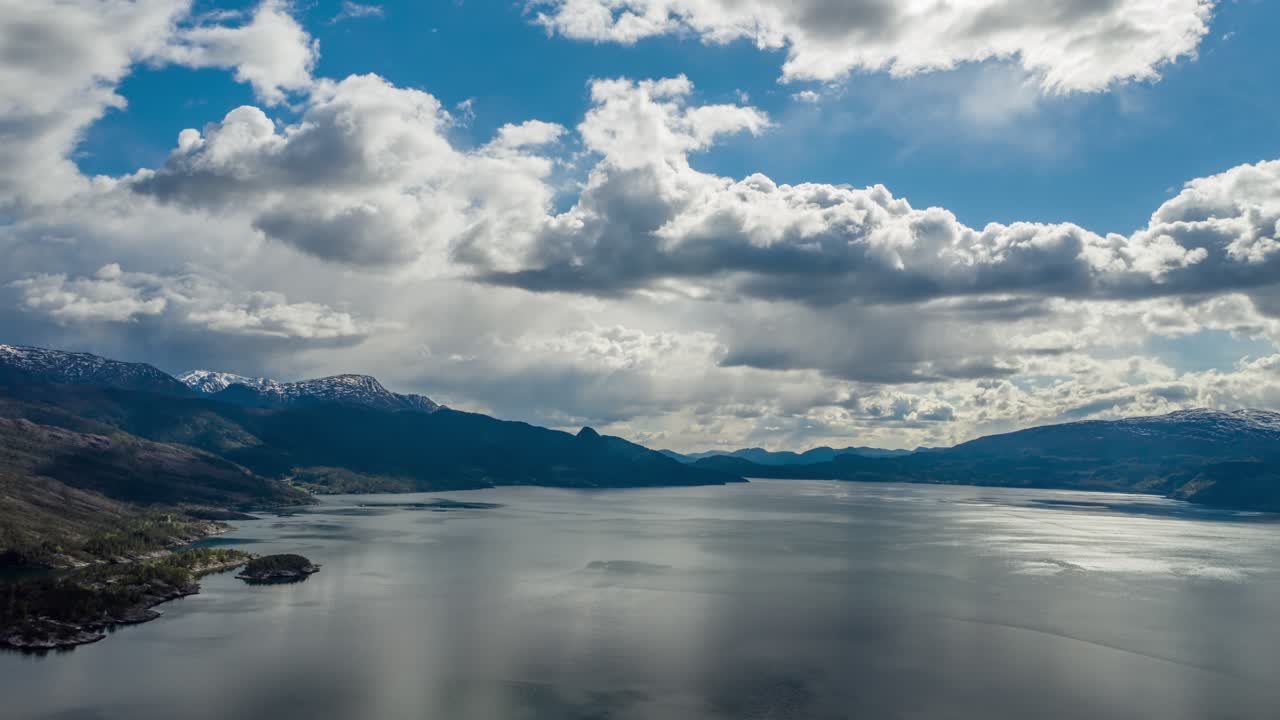 Thick, white-gray, stormy clouds whirling above the dark blue waters of the Hardanger fjord, Norway. Rays of sun are piercing through the clouds, lighting dark waters. Mountains tower on the horizon