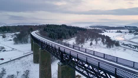 Sie-Fliegen-über-Das-Findhorn-Viadukt,-Während-Sich-Eine-Schafherde-Auf-Dem-Schneebedeckten-Feld-Versammelt