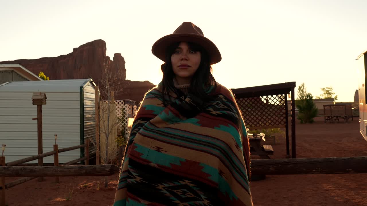 Mujer joven con sombrero y manta de pie en el desierto al atardecer