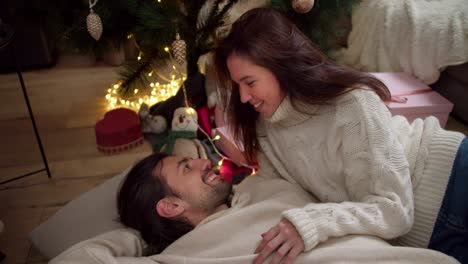 A-brunette-guy-is-lying-on-a-pillow-on-the-floor,-his-brunette-girlfriend-in-a-White-sweater-is-looking-at-him-and-hugs-him.-Near-the-New-Year-tree-and-gifts-in-a-room-decorated-for-the-New-Year-in-winter