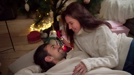 un chico moreno está tendido en una almohada en el suelo, su novia morena en un suéter blanco lo está mirando y lo abraza. cerca del árbol de año nuevo y regalos en una habitación decorada para el año nuevo en invierno