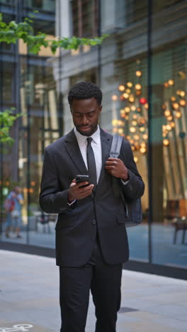 Vertical-Video-Shot-Of-Young-Businessman-Wearing-Suit-Using-Mobile-Phone-Walking-Outside-Past-Offices-In-The-Financial-District-Of-The-City-Of-London-UK-Shot-In-Real-Time