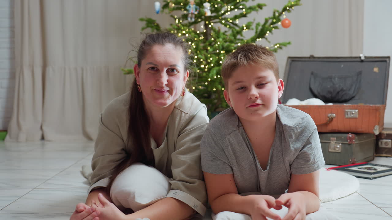 woman and son lying on white rug using white pillows smiling warmly christmas tree with lights in background cozy indoor setting ornaments scattered nearby joyful festive family bonding atmosphere