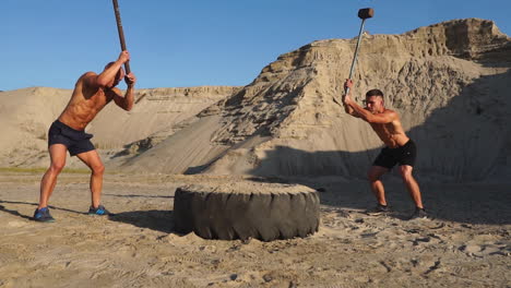 two powerful male athletes training together hit the wheel with a hammer at sunset in the desert. endurance training