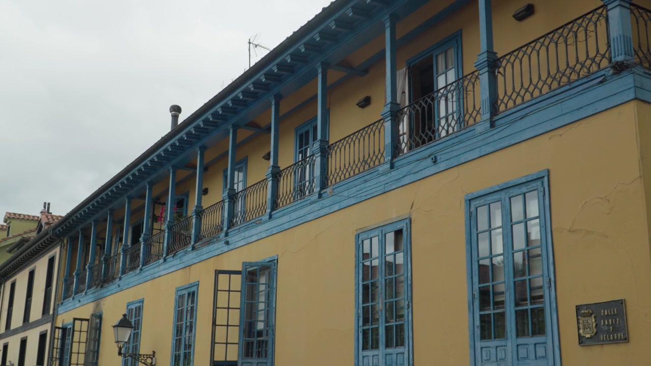 Vista fixa mostra uma fachada amarela de Oviedo com varandas azuis e janelas altas