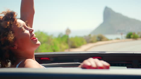 Portrait-Of-Woman-Standing-Up-Through-Sun-Roof-Of-Car-On-Road-Trip-With-Friends-Through-Countryside