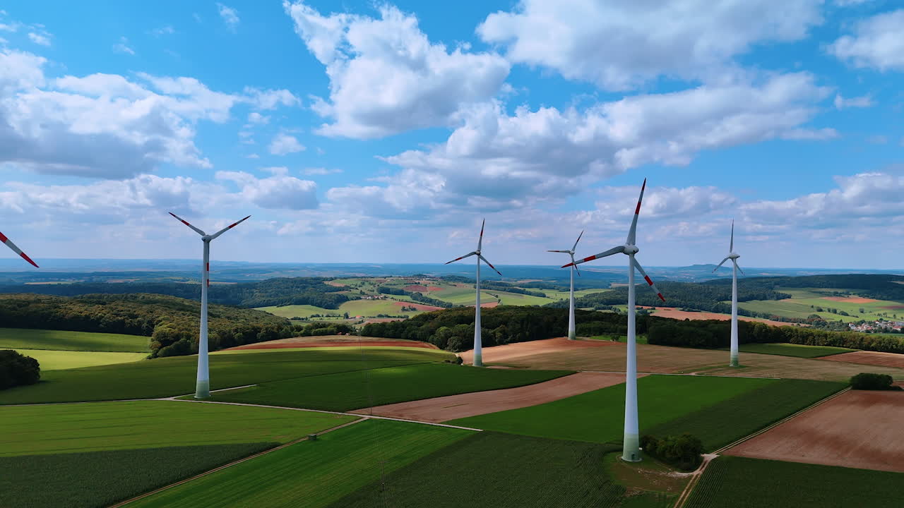 Wind turbines on green and brown fields under partly cloudy sky. Panoramic aerial shot of wind turbines generating energy above farmlands in a peaceful rural area
