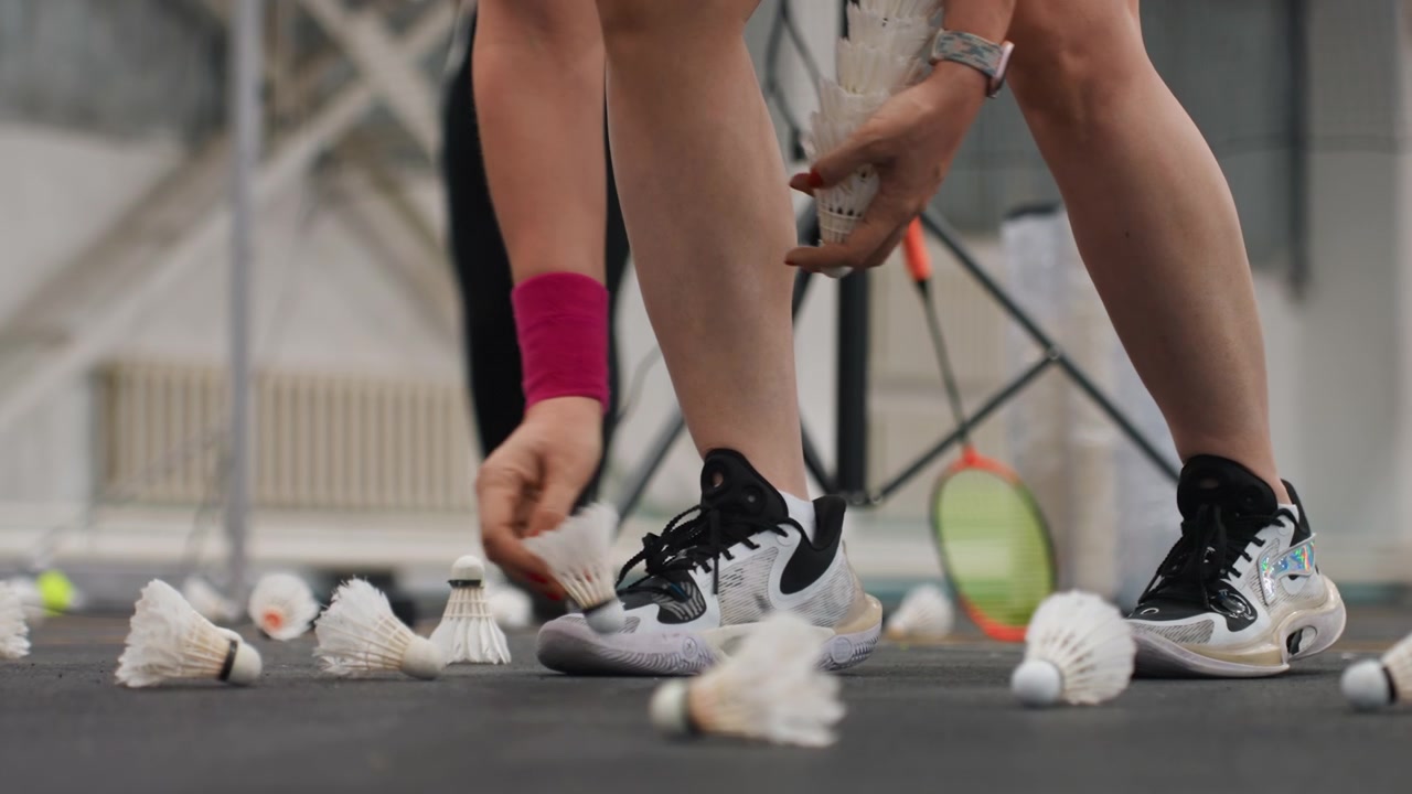 Volani sparsi su campo al coperto, giocatore si inginocchia per raccogliere volani bianchi con piume vicino a scarpe da ginnastica e racchetta, assistente regola il treppiede mentre l'allenatore osserva l'esercizio, primo piano su mani e piedi.