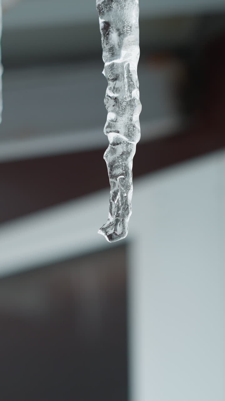 금속 지붕 가장자리의 클로즈업으로 긴 얼음이 녹고 물이 흘러내리며, 복잡한 얼어붙은 세부 사항과 흐릿한 배경에 형성되는 물방울을 강조합니다.