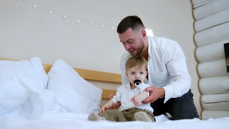 bearded man in white shirt stands behind his baby boy. father plays with his son clapping hands.