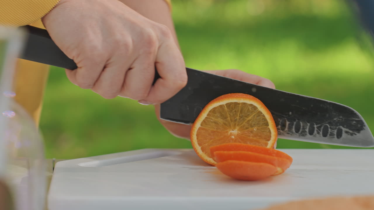 Mano che sistema fette d'arancia per un picnic in campeggio. Disposizione ordinata di fette su tagliere, luce solare calda e sfondo verde, brocca e coperta vicine, genitore che prepara uno spuntino di famiglia per un raduno all'aperto.