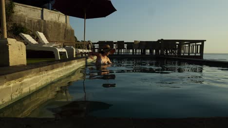 slow motion shot of a model getting into the infinity pool and swimming overlooking suluban beach during sunset at golden hour