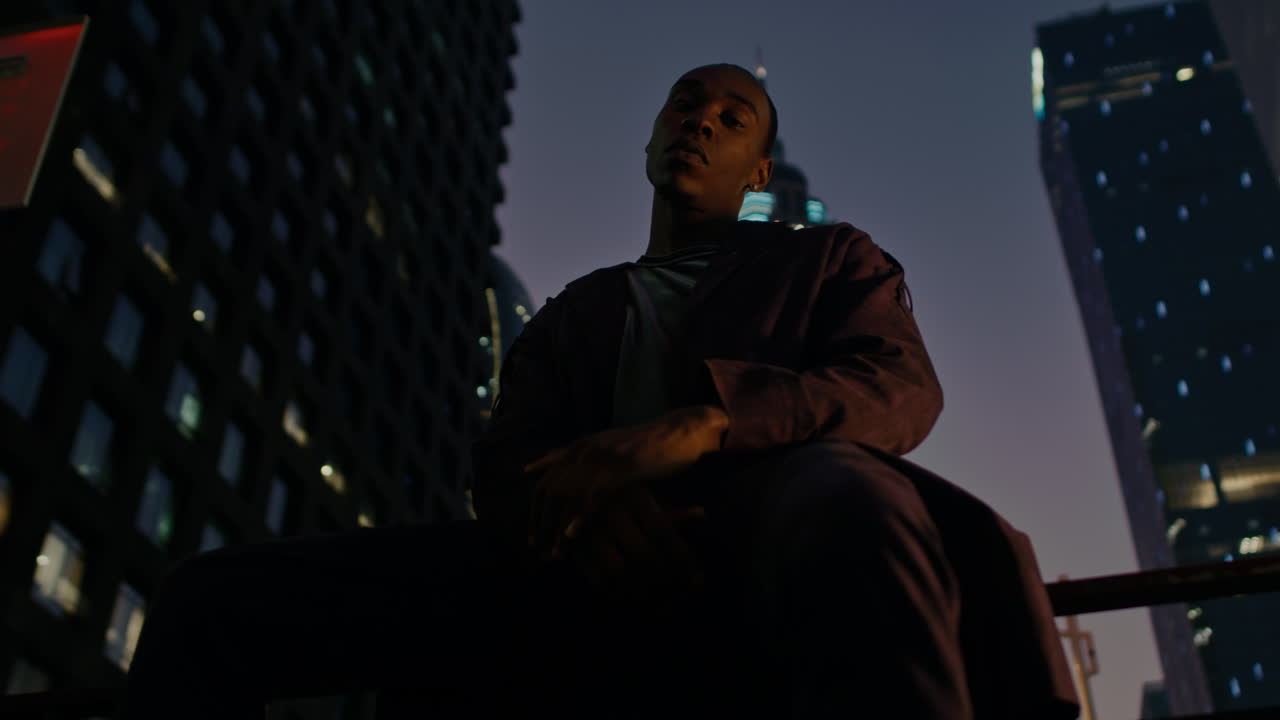 Afro American model sitting in front of camera in smart city with modern skyscraper building illuminated at night in background