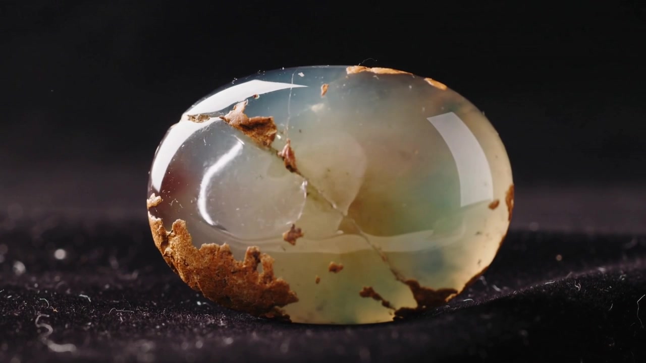 Lumière changeante faisant tourner une gemme cabochon sur un tissu sombre, montrant une fine fissure et une matrice brune.