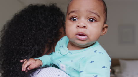 amorosa madre afroamericana con pijama acurrucando al bebé hijo en el dormitorio en casa