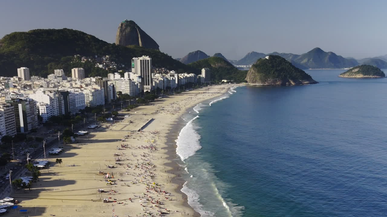 Drone flying over Copacabana Beach, people enjoying beach, swimming in blue water, white waves, city skyscrapers, with Sugar Loaf Mountain and islands in background