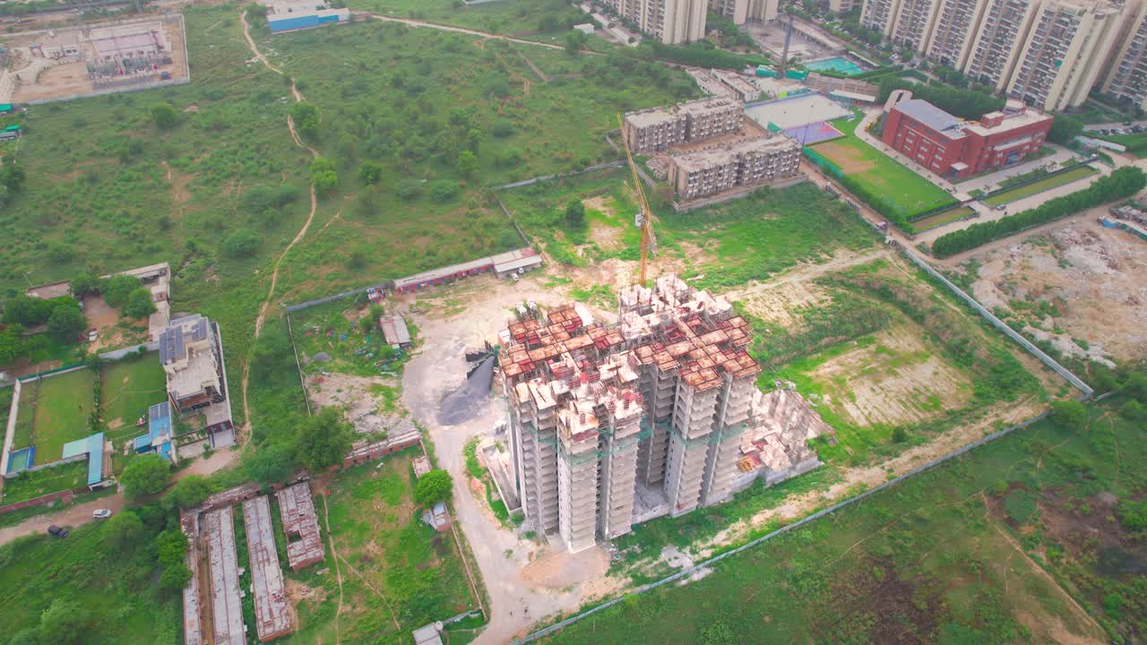 Aerial drone shot flying backwards revealing under construction building surrounded by green fields showing urbanization, development in real estate and growing housing in cities