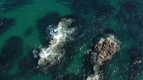 Aerial-top-down-view-of-ocean-waves-crashing-into-small-rocks-in-shallow-water