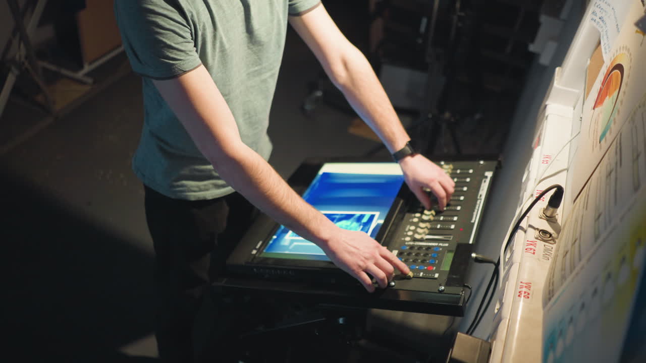 Man adjusting monitor, focused on electronic control panel in studio with detailed papers and settings visible in background. Low-light indoor setting with blurred equipment and soft light accentuating scene