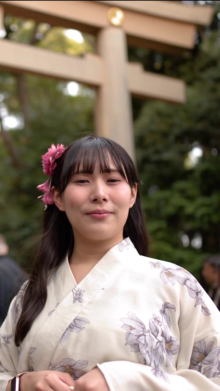 En smilende japansk kvinde iført en blomstret yukata står foran en stor træ-torii-port.