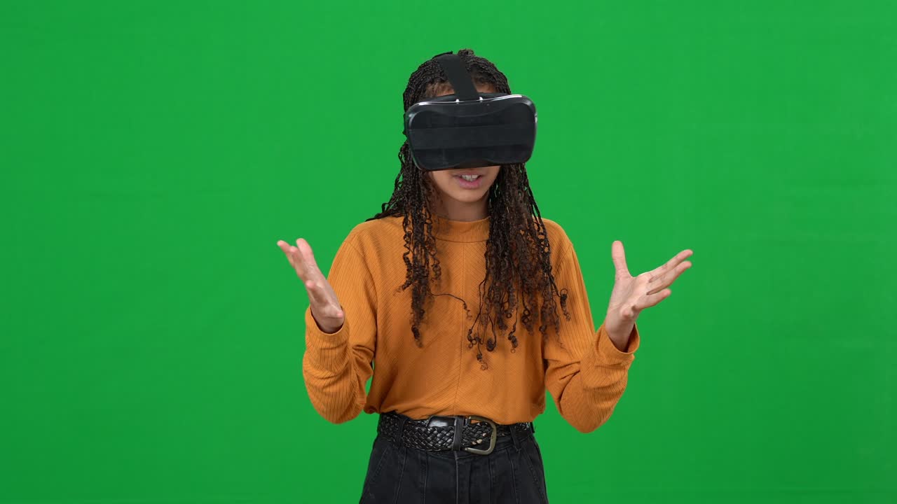 Absorbed African American teenage girl in VR headset pointing around turning on green screen. Portrait of excited female gamer enjoying augmented reality at chromakey background.